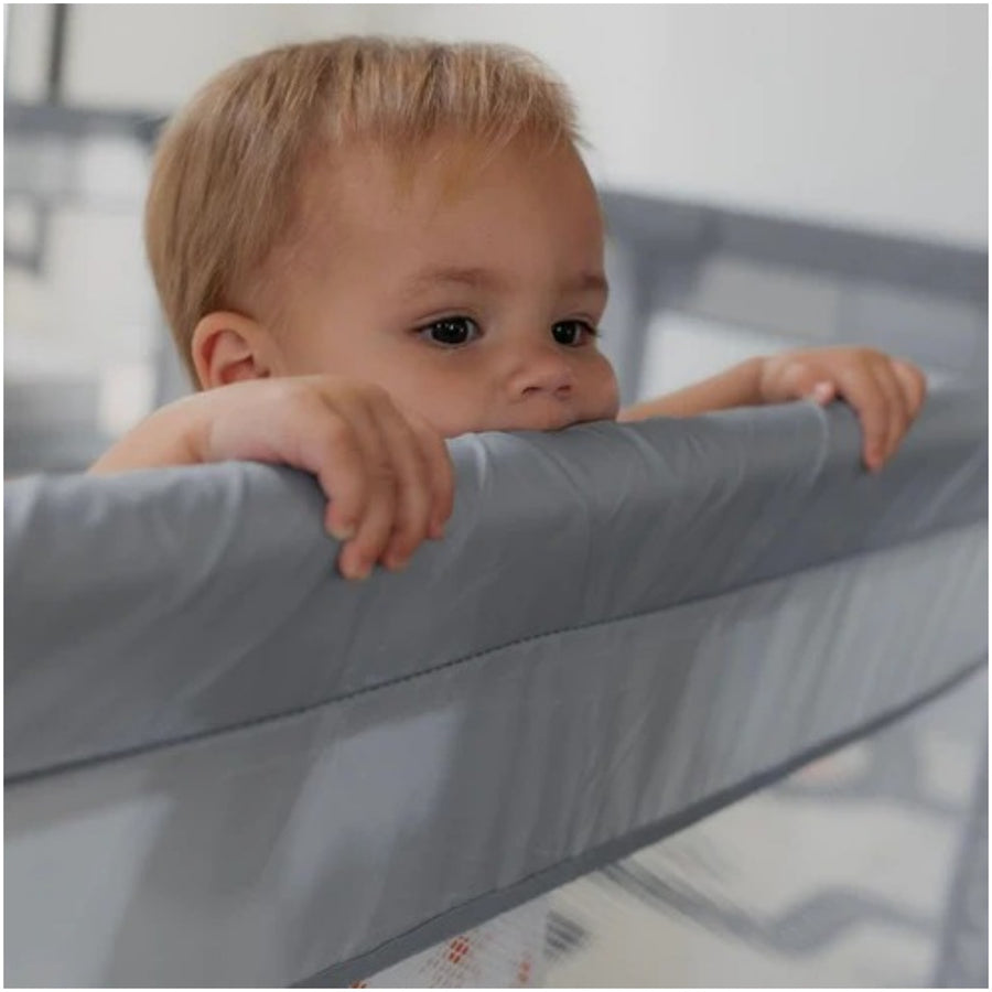 Baby standing and holding onto the padded top rail of the play yard