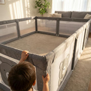 Small hands gripping the padded top rail of the large grey play yard